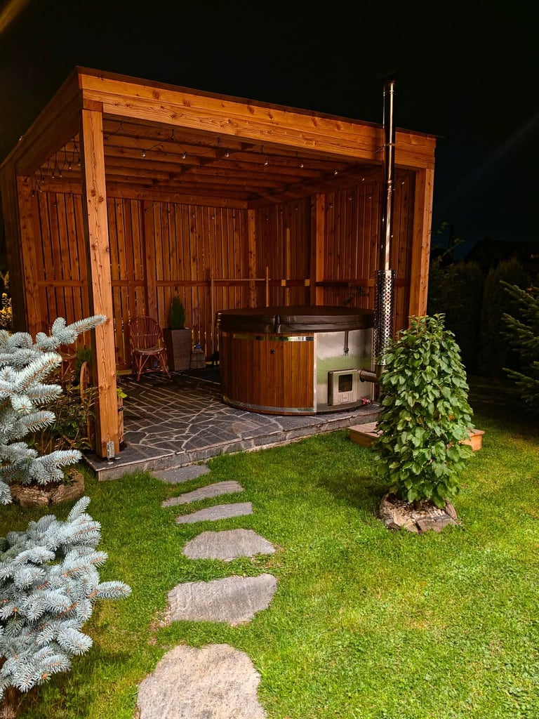 Drewniana altana ogrodowa z balią jacuzzi nocą, otoczona zielenią i kamiennymi płytami